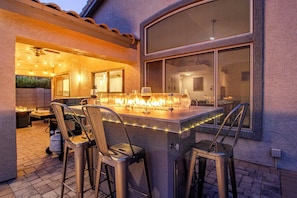 An outdoor bar area with a fire feature, bar stools, and a view of the interior through a window. - Gold Canyon Superstition Foothills Retreat w/ Pool (Gold Canyon)