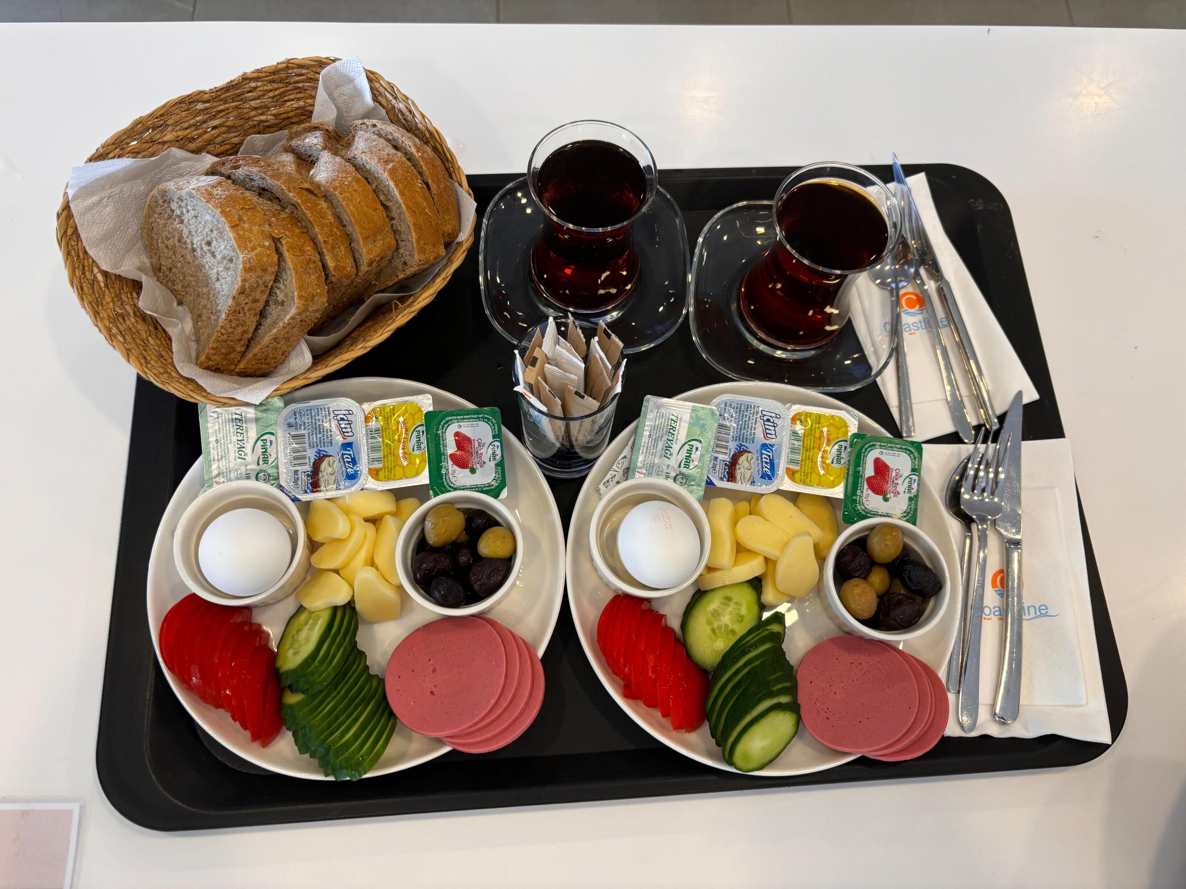 Een dienblad met sneetjes brood, twee glazen rode wijn, en schotels met kaas, olijven, komkommers en ham.