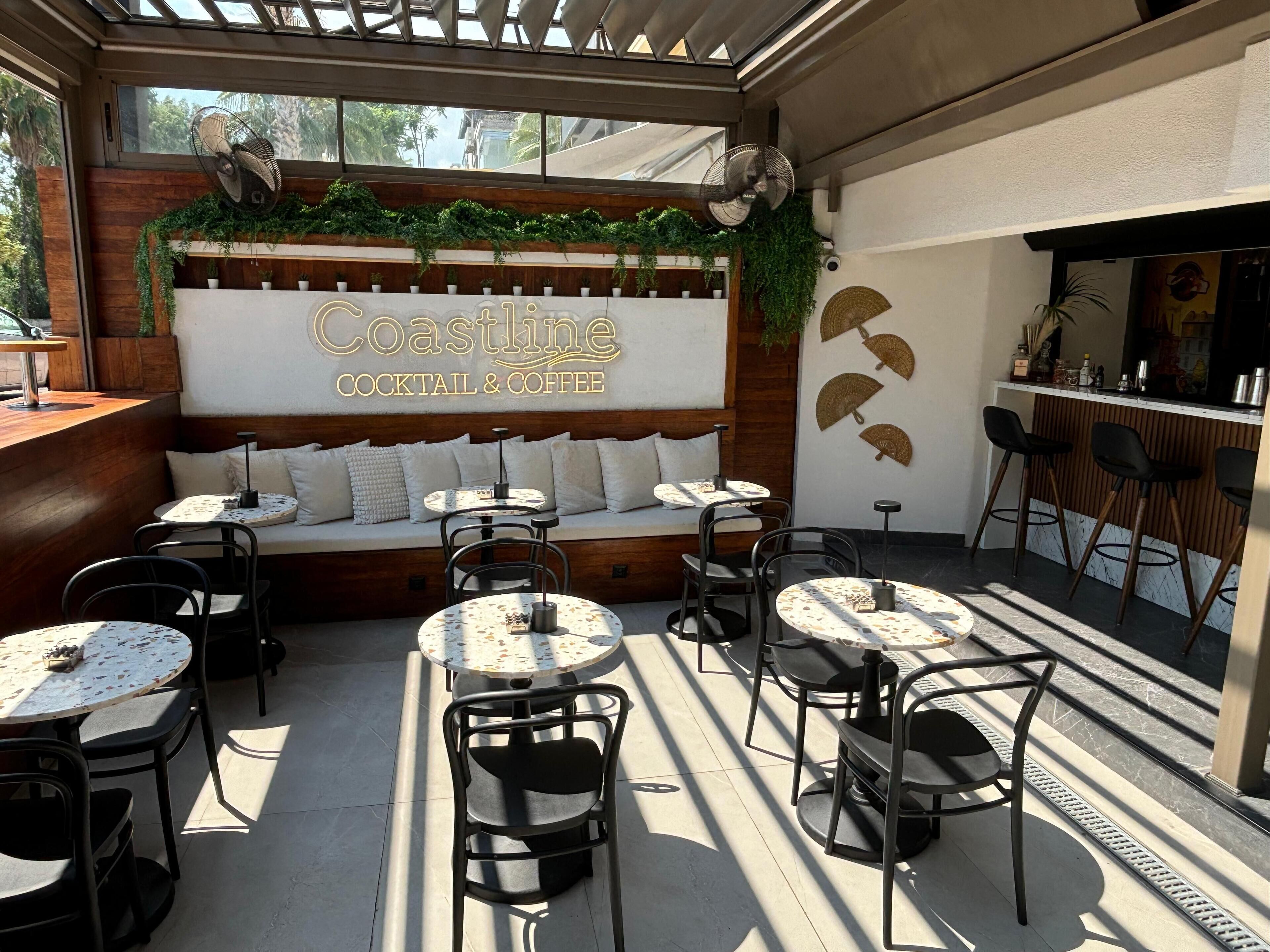 An outdoor seating area with round tables and chairs, a wooden bench, and a wall with 'Coastline Cocktail & Coffee' signage.