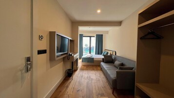 Una habitación de hotel moderna con piso de madera, un sofá gris, una cama con cabecera azul, un televisor montado en la pared y un balcón con cortinas.