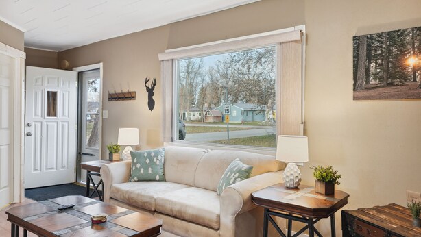 A living room with a beige sofa, two side tables, a wooden coffee table, a door, a window with a view of houses, and a mounted deer head on the wall. - Choteau Vacation Rental w/ Yard: Walk to Downtown! (Choteau)