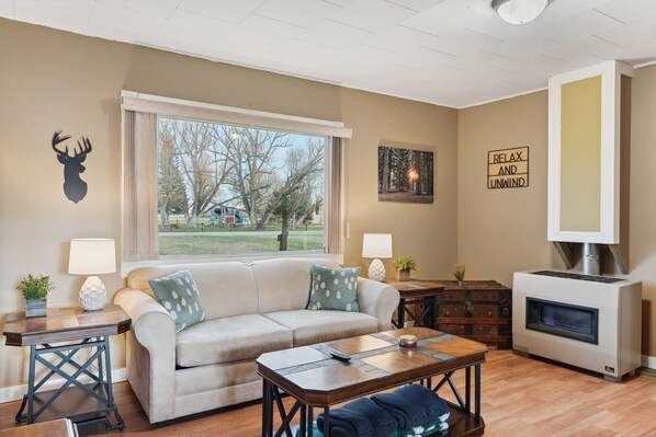 A living room with a beige sofa, a wooden coffee table, a fireplace, and a view of a park through a large window.