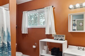 A bathroom with a white toilet, a shelf with towels and a sign, a window with white curtains, and a mirror with lights above.