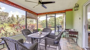 Une terrasse couverte, aménagée avec des meubles en osier, un ventilateur de plafond et offrant une vue sur le jardin.