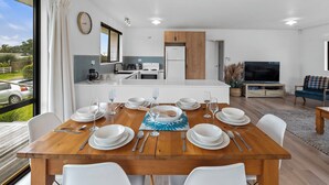 Una cocina moderna con electrodomésticos blancos, un comedor con una mesa de madera dispuesta para cuatro personas, y una sala de estar con televisor y sofá.