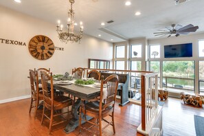 A dining area with a wooden table, chairs, and a chandelier. There is a clock on the wall and a TV mounted on the wall. - Lakefront Townhome in Pottsboro: Pets Welcome! (Pottsboro)