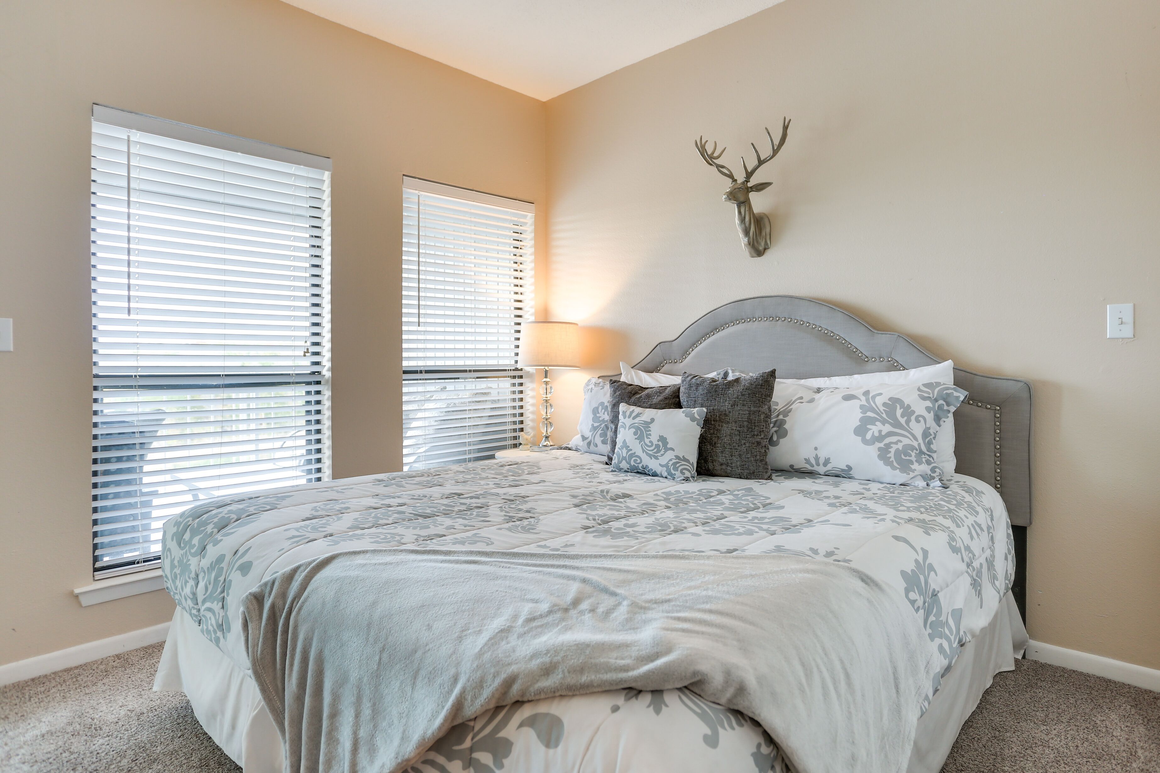 A bedroom with a large bed, two windows with blinds, a mounted deer head decoration, and a bedside lamp.