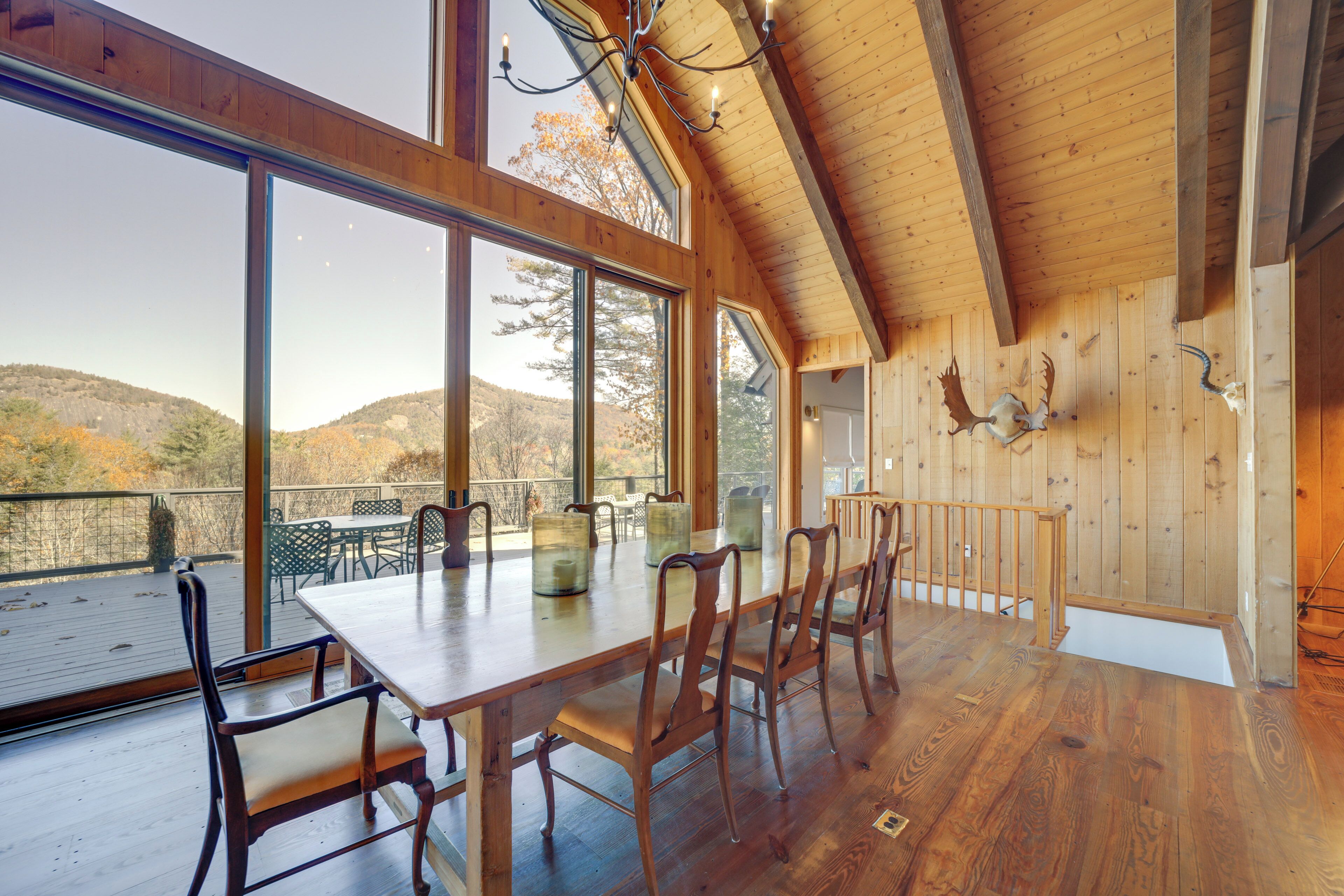A wooden interior with a large dining table and chairs, large windows offering a view of the outdoors, and a wooden ceiling with beams.