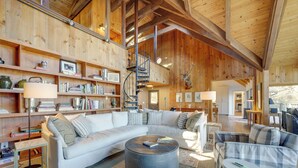A spacious living area with a large white sectional sofa, a round coffee table, and a built-in bookshelf. A spiral staircase leads to an upper level. - Mountain-view Cashiers Home: Near High Hampton!- (Cashiers)