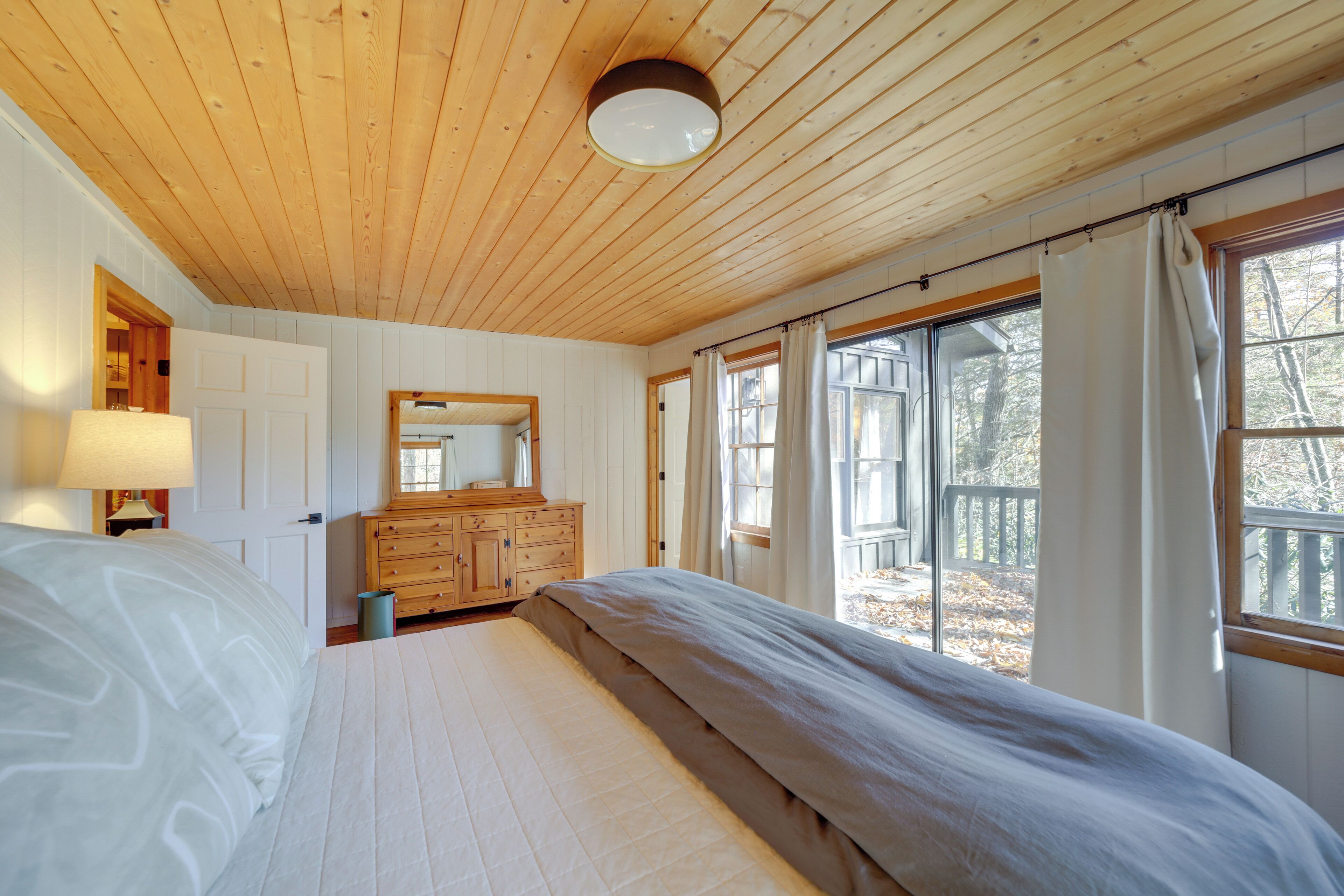 A bedroom with a wooden ceiling, a large bed, a dresser, a mirror, and a window with curtains.