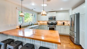 A modern kitchen with white cabinets, stainless steel appliances, a wooden island with bar stools, pendant lighting, and a large window providing natural light. - Lakefront Edgerton Cottage w/ Deck & Grill! (Edgerton)