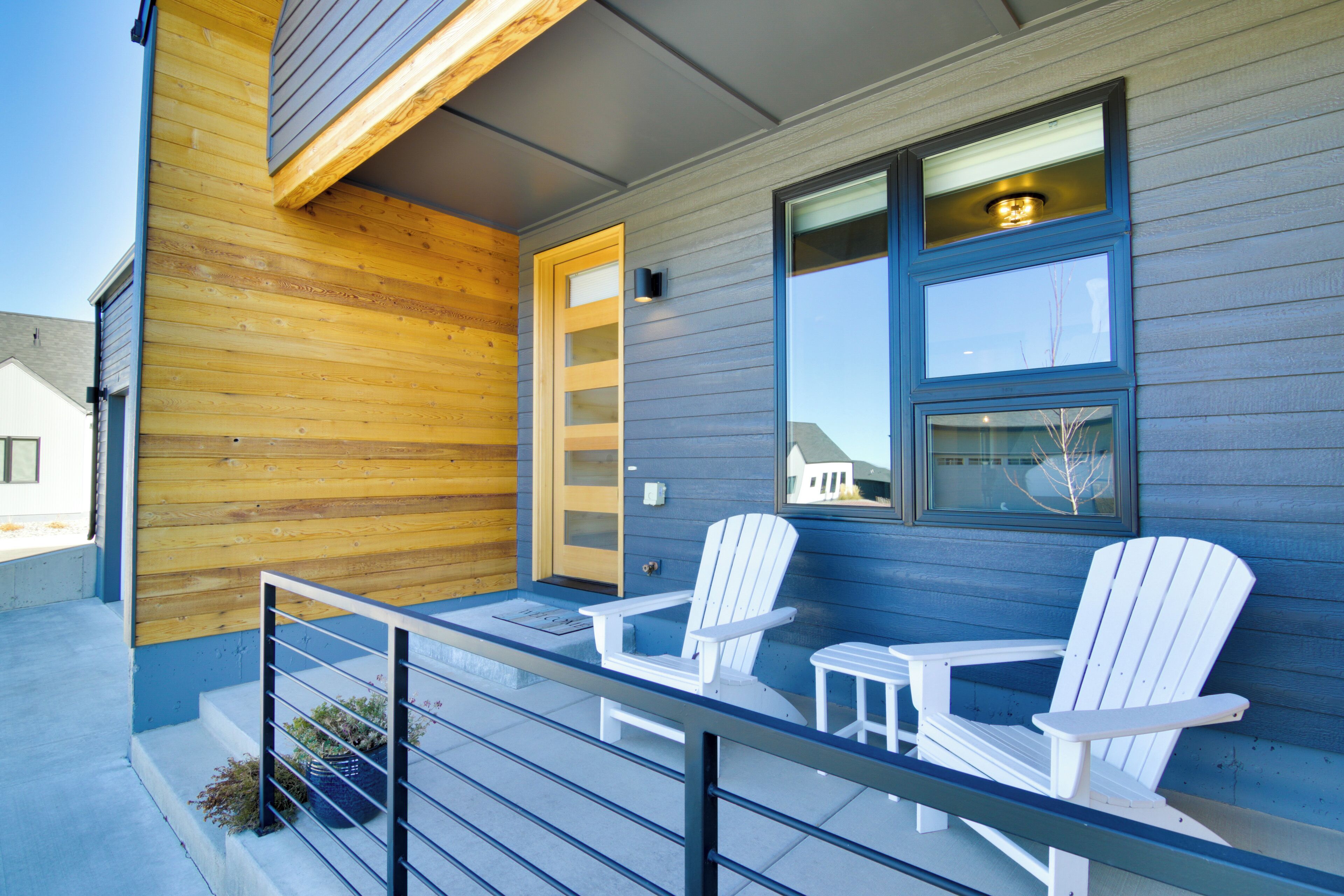 A wooden exterior with a blue door and windows, two white Adirondack chairs, and a small table on a balcony.