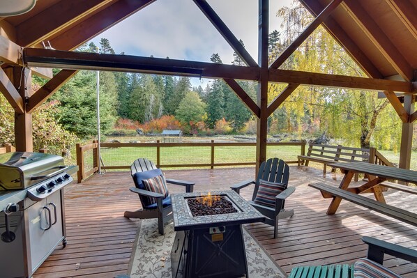 Une terrasse extérieure équipée d’un barbecue, d’un foyer et d’un espace salon, offrant une vue sur un paysage boisé.