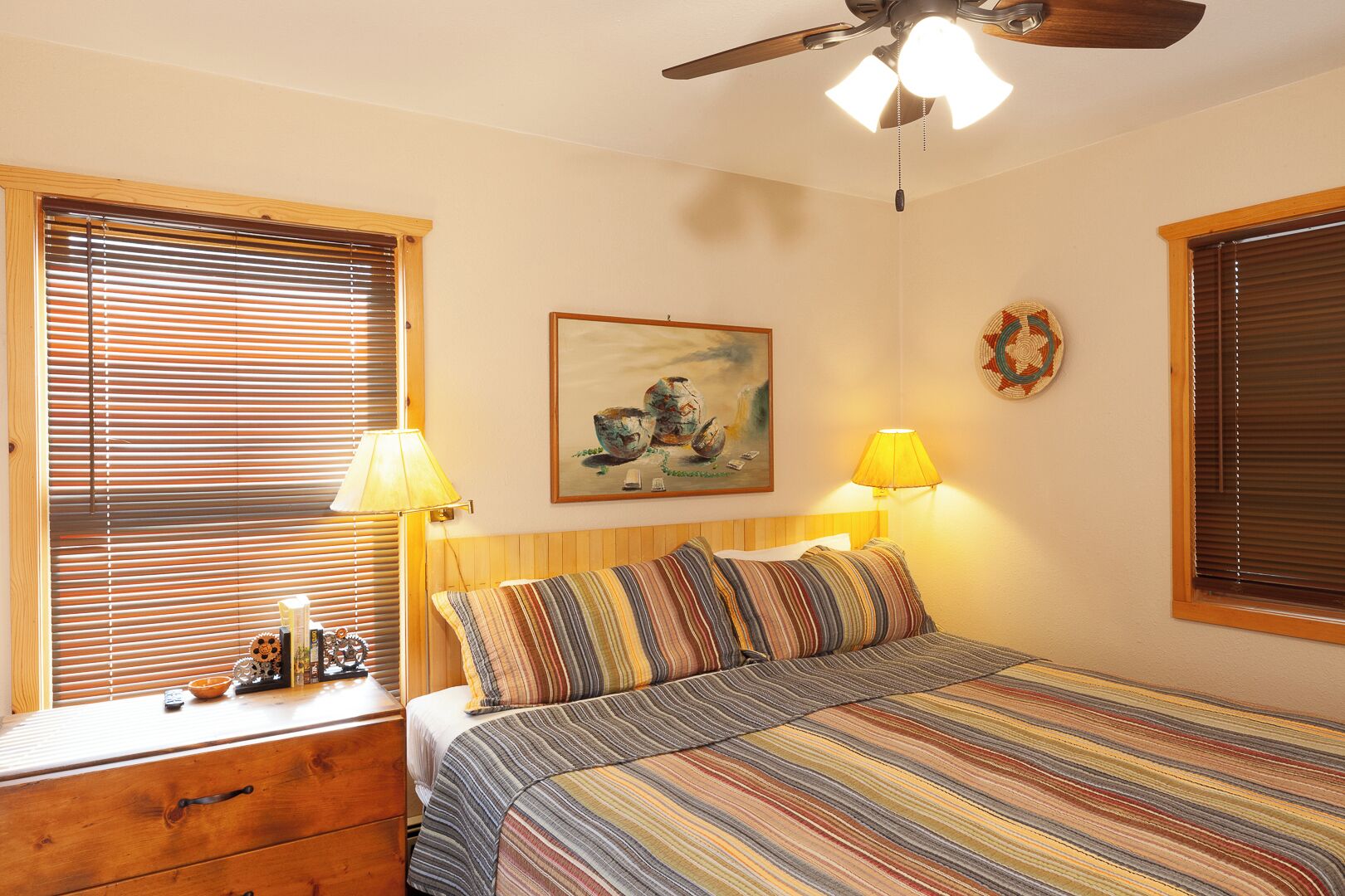 Ein Schlafzimmer mit einem Bett, zwei Nachttischlampen, einem Deckenventilator, einem Bild an der Wand und einem Fenster mit Jalousien.