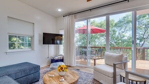 A modern living room with a large TV, a wooden coffee table with a vase of sunflowers, a grey sofa, and a beige armchair. - Jonestown Retreat w/ Deck: 6 Mi to Lake Travis! (Leander)