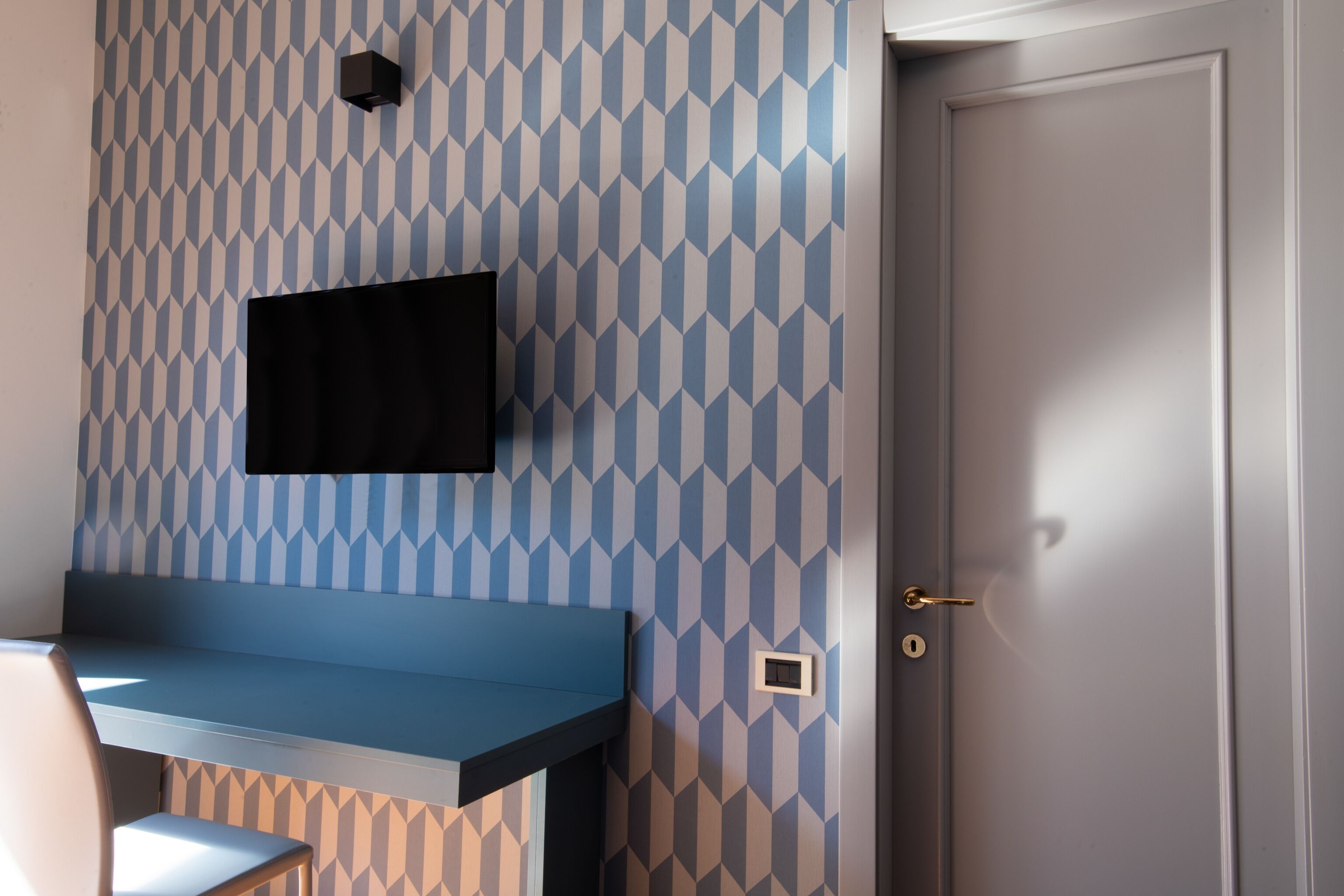 Une pièce avec un mur orné d’un motif géométrique, un téléviseur à écran plat fixé au mur, un banc bleu et une porte dotée d’un trou de serrure.