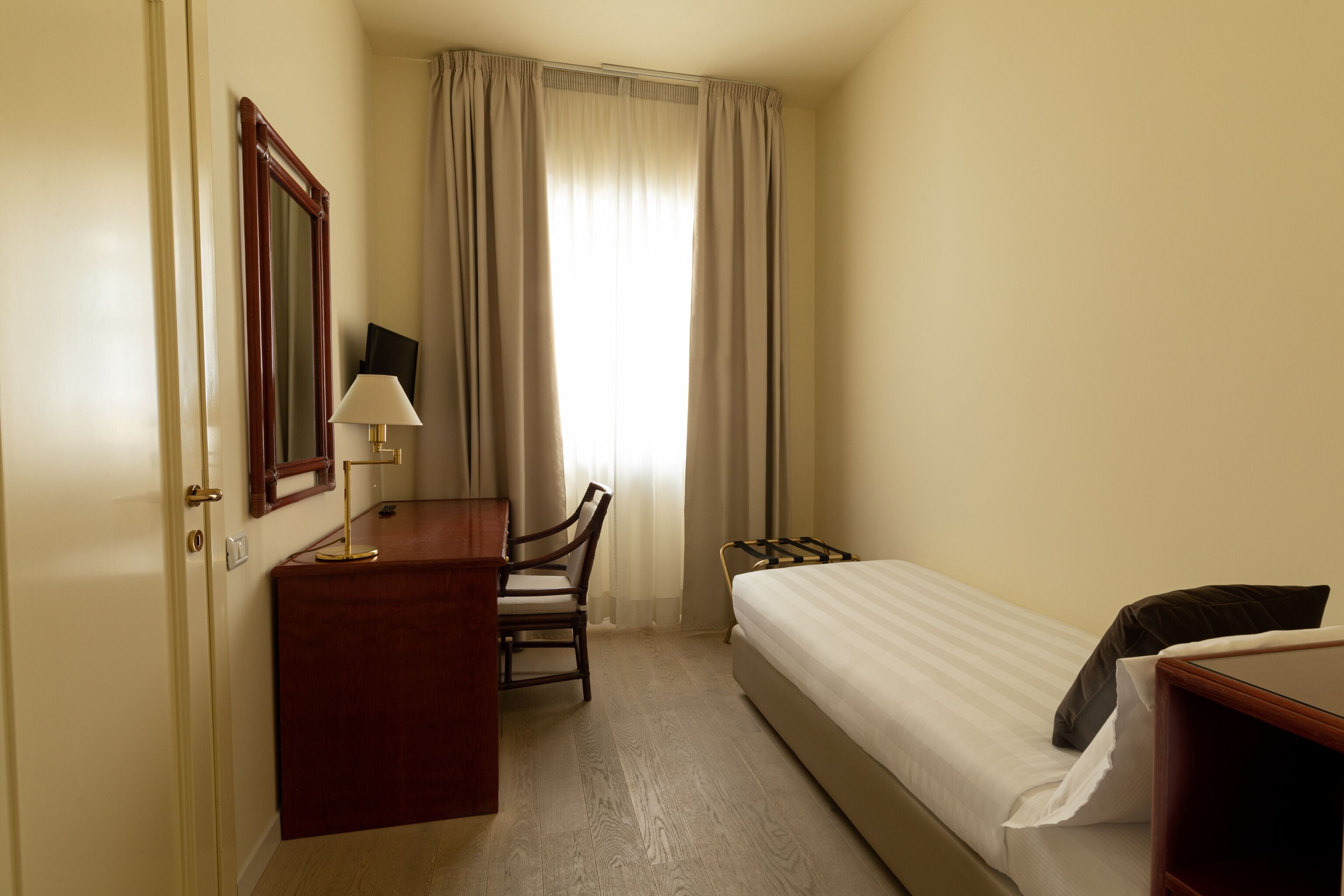 Habitación de hotel con cama, escritorio con silla, lámpara y un ventanal con cortinas.