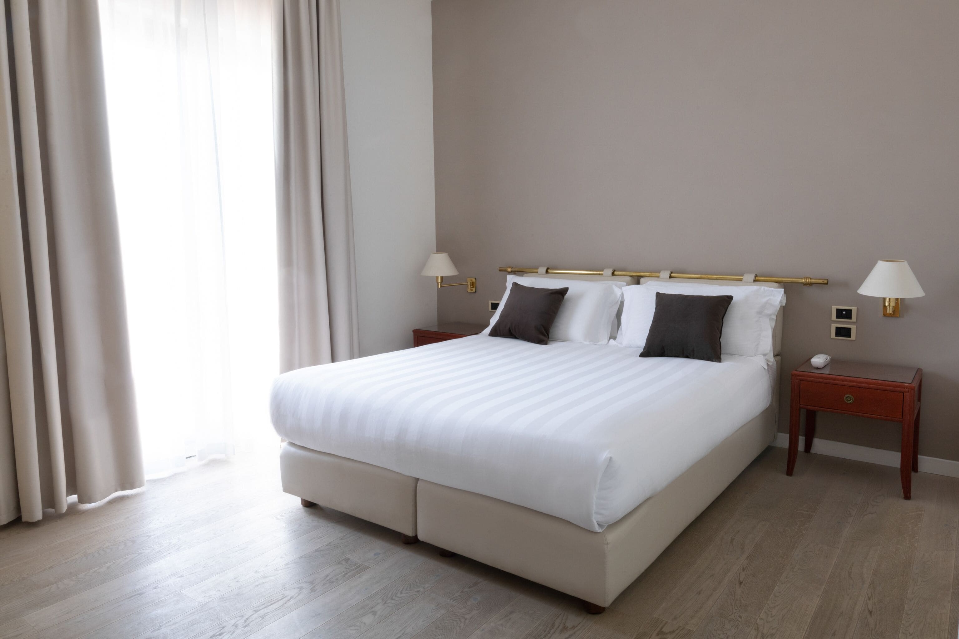 A neatly made bed with white linens and two pillows, a wooden nightstand with a lamp, and a wooden floor.