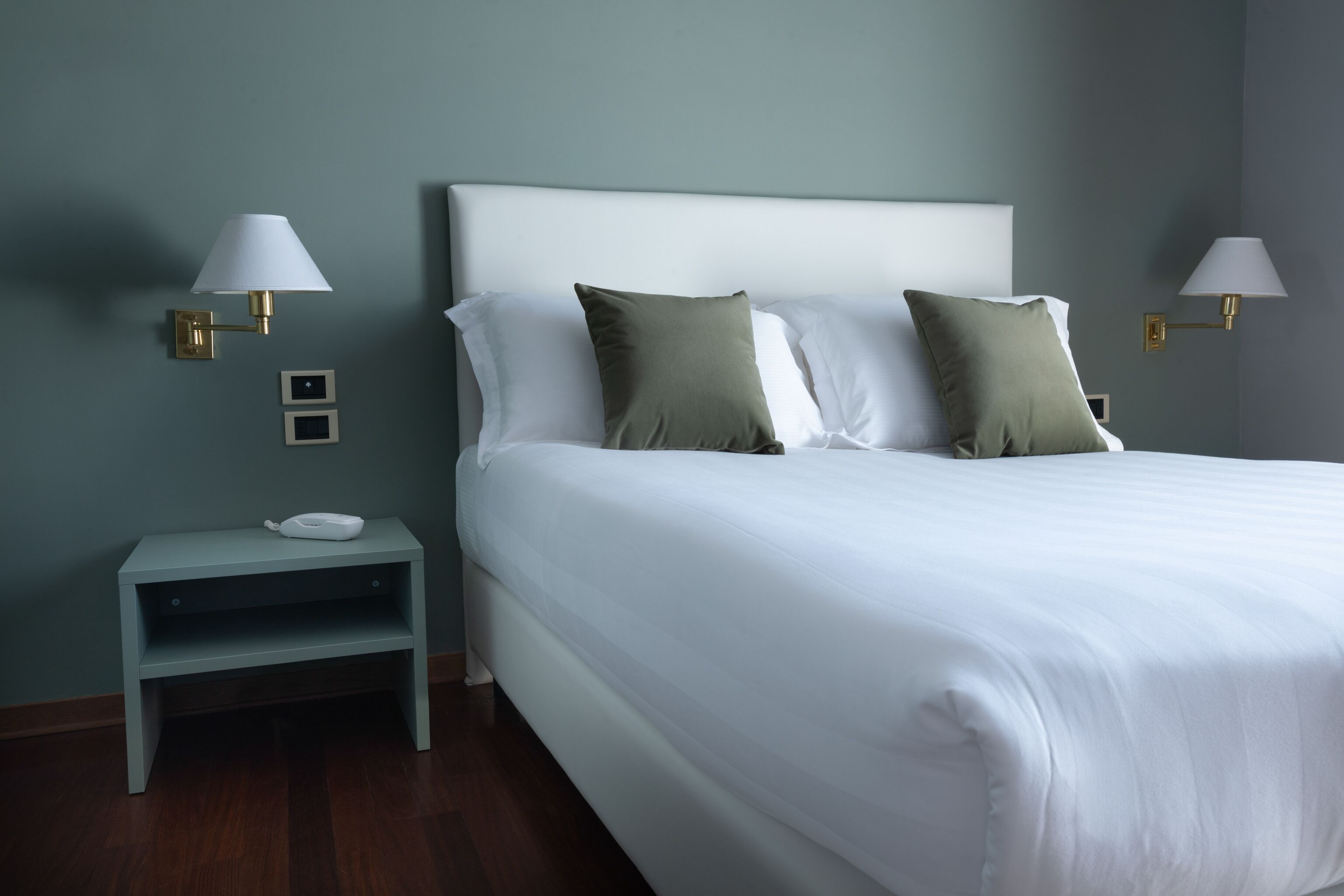 Una cama bien hecha con sábanas blancas, dos almohadas y una mesita de noche con una lámpara.
