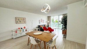 A modern dining area with a wooden table and white chairs, a white console table with decorative items, and a large window with a view of greenery. - Beachfront Apt | Tides Hollywood 5th Floor + Pool (Hollywood)