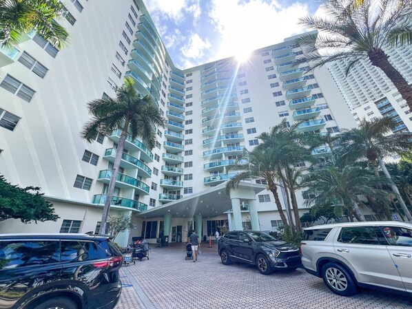 A modern high-rise building with multiple balconies, a paved entrance area, and parked cars.