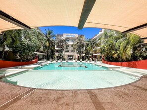 Area piscina con vasca azzurra e trasparente, circondata da palme e un edificio con balconi sullo sfondo.