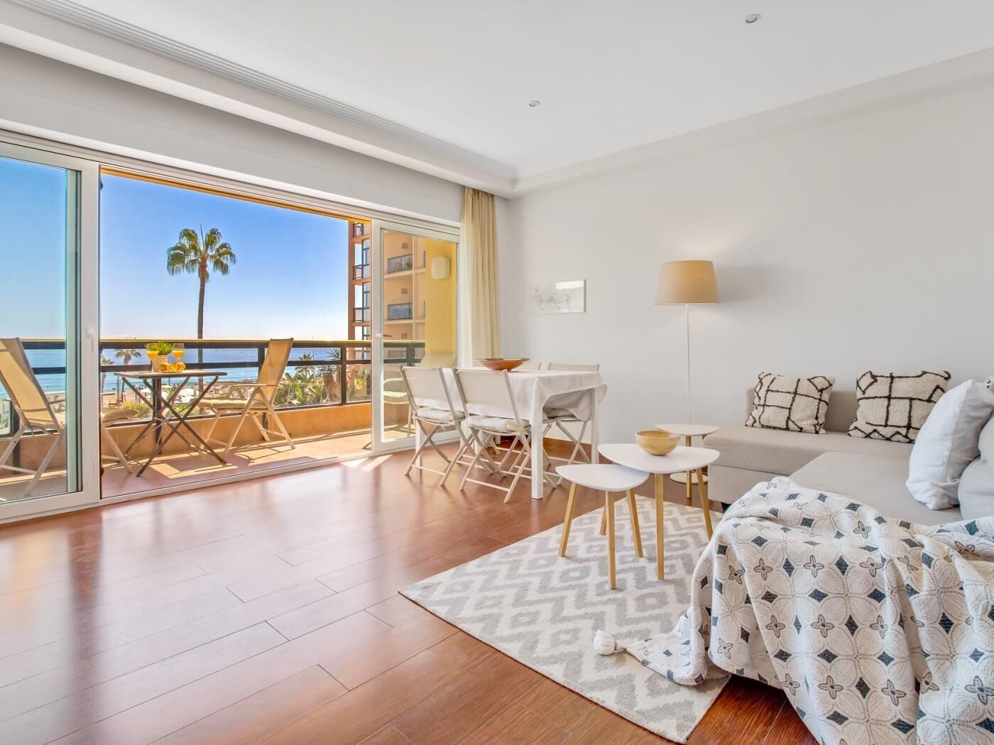A spacious balcony with a view of the sea, a dining area with white furniture, and a bed with patterned bedding.