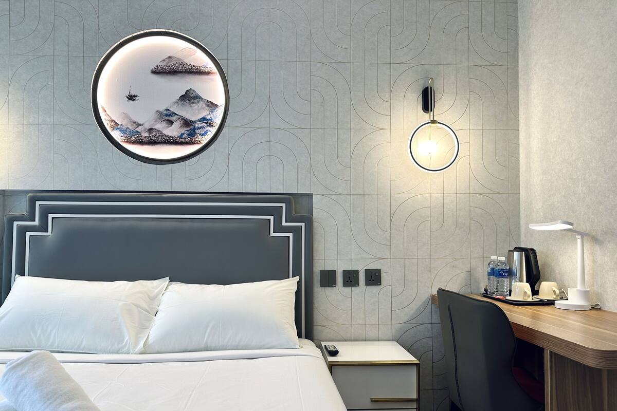 A hotel room with a bed, a desk, a chair, a round wall decoration, and a wall-mounted light fixture.