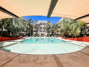 A pool area with a clear blue pool, surrounded by palm trees and a building with balconies in the background. - Sabbia 4310 - 3 Bedrooms All-in Suite (Playa del Carmen)