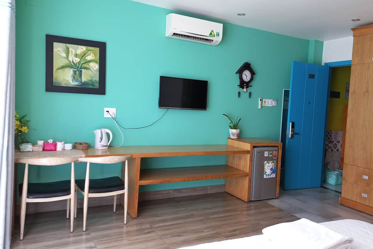 A room with teal walls, a wooden desk with a chair, a small refrigerator, a clock, and a TV mounted on the wall.