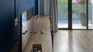A room with a blue wall, a ceiling fan, a large window with curtains, a wooden table, and a balcony with a potted plant. - Capybara Hotel (Ho Chi Minh City)