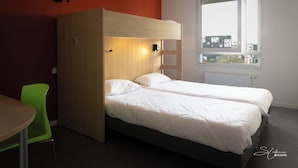 Una habitación con dos camas, una litera de madera, una silla verde, una mesa y una ventana con vista a la ciudad.