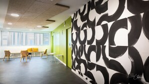 Un espacio de oficina moderno con un largo pasillo, una zona de estar con sillas y mesas, y una gran obra de arte abstracto en la pared.