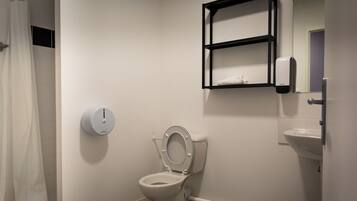 A white toilet with a closed lid, a wall-mounted soap dispenser, and a small shelf above the toilet.