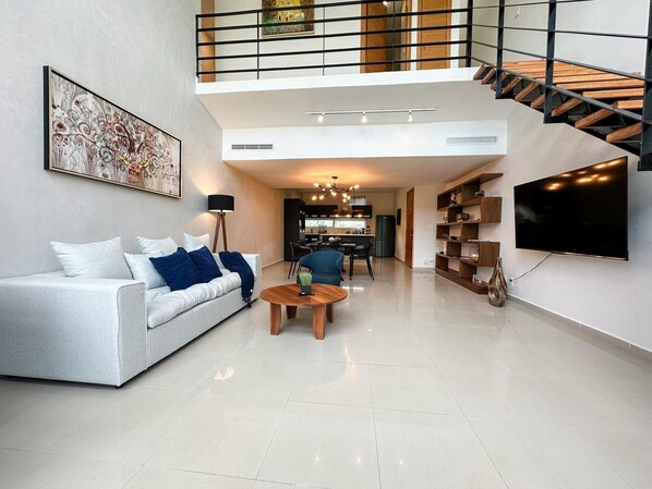 Un moderno salón con sofá, mesa de centro, televisor, escalera y cocina al fondo.
