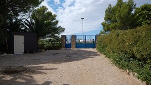 Une allée de gravier mène à une entrée fermée par un portail bleu, le tout étant entouré de verdure et bordé, sur la gauche, d’un petit bâtiment.