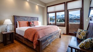 A bedroom with a wooden bed, a nightstand, a lamp, a view of mountains through large windows, and a small table with a chair.