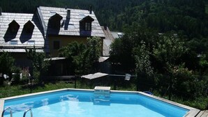 A swimming pool with a wooden deck and metal ladder, surrounded by lush greenery and a house with a sloped roof. - Cozy Studio with Wi-Fi, BBQ, Summer Pool, Near Ski Pistes & Free Village Shuttle (Saint-Chaffrey)