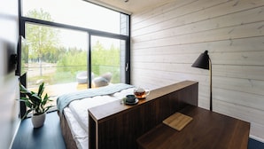 A modern bedroom with a large window, a bedside table with a teapot, and a view of greenery outside. - PihlasResort (Joroinen)
