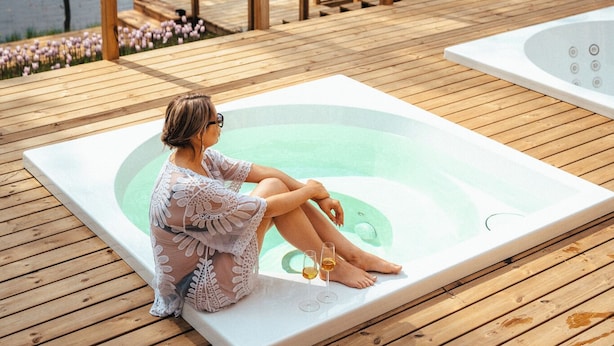 A person relaxing on a wooden deck by a lake, with a hot tub and a table with chairs. - PihlasResort (Joroinen)