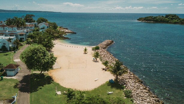 An aerial view of a beach with a rocky shoreline, a clear blue sea, and a row of houses along the water's edge. - Palm Point - 2/2 Townhouse Steps from the Beach (Negril)