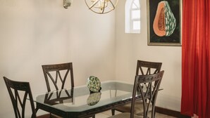 A dining area with a glass table, four wooden chairs, a decorative light fixture, a framed fruit illustration, and a red curtain. - Palm Point - 2/2 Townhouse Steps from the Beach (Negril)