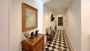 Um corredor com chão xadrez, um banco de madeira, um espelho com moldura dourada e uma porta com padrão geométrico.