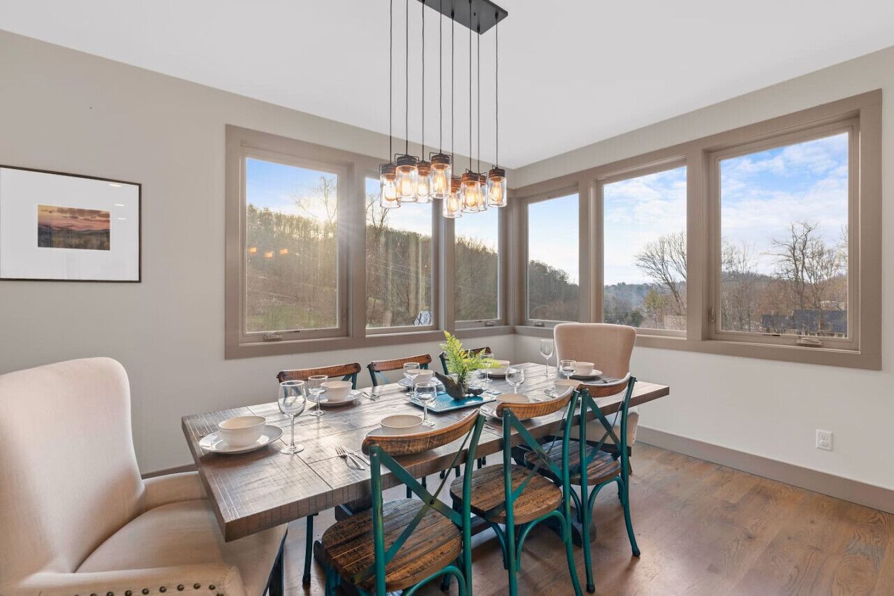 A dining area with a wooden table set for six, surrounded by chairs, large windows with a view of trees, and a hanging light fixture.