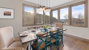 Un comedor con una mesa de madera dispuesta para seis personas, sillas alrededor, ventanas amplias con vistas a árboles y una lámpara colgante.