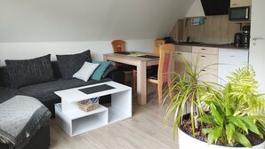Un espacio habitable compacto con un sofá, una mesita, una silla, una planta en maceta y una kitchenette.