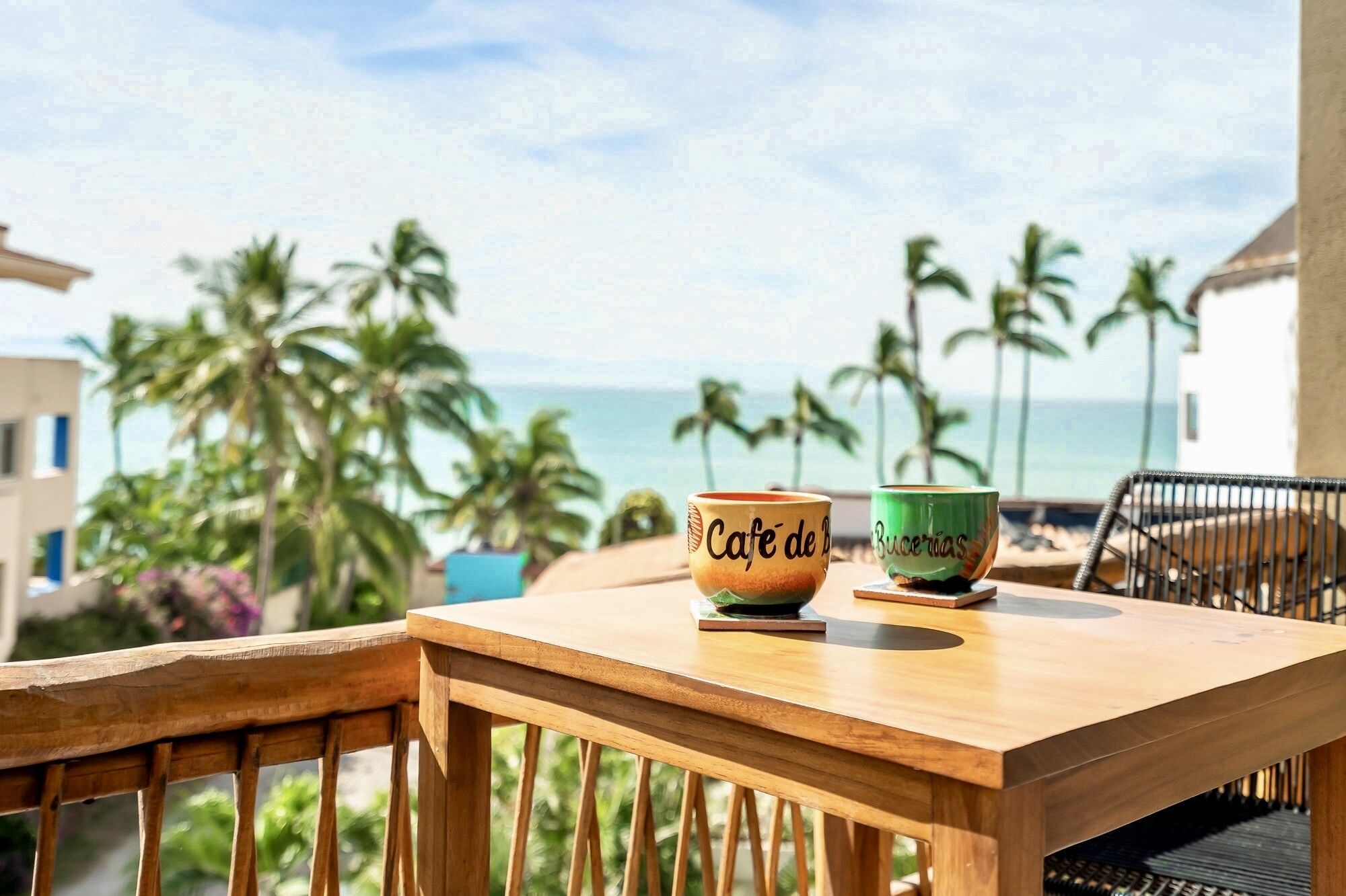 Una mesa de madera con dos tazas de cerámica que dicen "Cafe de B" y "Buccinas", en un balcón con vista a una playa con palmeras y el océano.