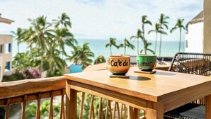 A wooden table with two ceramic cups labeled "Cafe de B" and "Buccinas" on a balcony overlooking a beach with palm trees and the ocean. - Casa Kaia, Ocean-View Boho-Chic, 2bd/2ba Condo in Golden Zone (Bucerías)