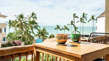 Una mesa de madera con dos tazas de cerámica que dicen "Cafe de B" y "Buccinas", en un balcón con vista a una playa con palmeras y el océano.