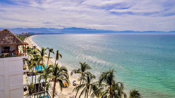 A beach with clear turquoise water, palm trees, and a thatched-roof building. - Casa Kaia, Ocean-View Boho-Chic, 2bd/2ba Condo in Golden Zone (Bucerías)
